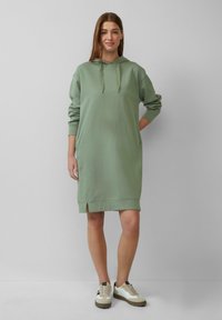 Grünes Hoodie-Kleid mit langen Ärmeln, Fronttasche und seitlichen Schlitzen am Saum. Kombiniert mit weißen und beigen Sneakern. Glatter Stoff. 