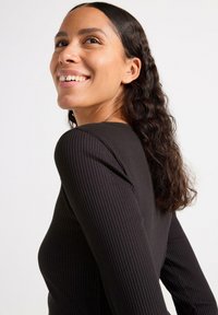 Top lungo a maniche nere a coste con scollatura rotonda, caratterizzato da cuciture senza soluzione di continuità e una silhouette aderente. I capelli sono mossi e di lunghezza media.
