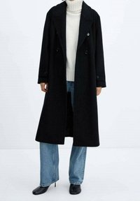 Cappotto in lana nera con design doppiopetto, ampi rever e dettagli con bottoni, indossato sopra un dolcevita bianco e jeans blu.