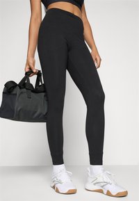 Person som har på seg svarte leggings, hvite treningesko, hvite sokker, holder en svart treningsduffelbag med den ene hånden, og står på en hvit overflate.
