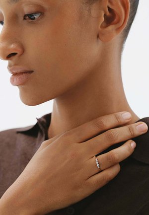 Mujer de piel morena que lleva un anillo de plata con cinco pequeños diamantes en el dedo anular, con la mano apoyada en el cuello, vistiendo una camisa marrón oscuro.