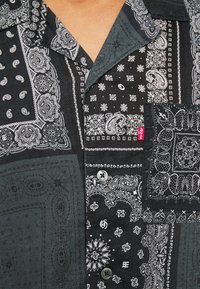 Camicetta nera in tessuto con motivi bianchi a paisley e floreali, chiusura con bottoni e taschino; design testurizzato con motivi differenti.