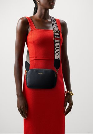 Žena v červených šatech bez rukávů s černou kabelkou přes rameno s širokým páskem s logem a zlatými detaily na zipu.