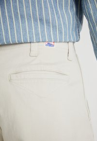 Pantaloni beige con taglio dritto, dotati di una tasca posteriore con un design diagonale e un piccolo etichetta del logo sopra la vita.