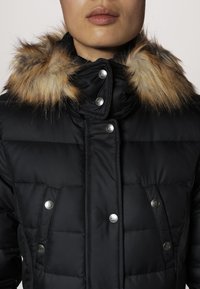 Chaqueta acolchada negra con un cuello de piel sintética, botones a presión y un diseño de acolchado horizontal. La tela es suave con un acabado mate.