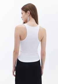 Witte geribbelde tanktop met een aansluitend ontwerp, dikke banden en een ronde rug, gecombineerd met een vloeiende zwarte rok. Zilveren statementoorbellen zichtbaar.