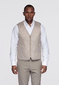 Chaleco acolchado beige con cierre frontal de cremallera, bolsillos laterales y diseño sin mangas, llevado sobre una camisa blanca con cuello y pantalones gris claro.