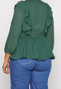 Blouse verte texturée avec détails à volants, portée avec un jean en denim bleu, vue de dos, de la taille aux épaules visibles.