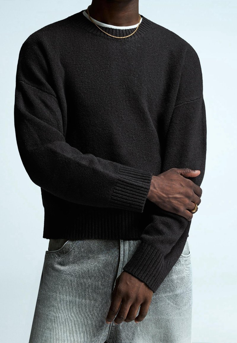 Personne portant un pull texturé noir par-dessus une chemise blanche, un jean gris clair, un collier chaîne en or et des bagues en or à la main gauche.