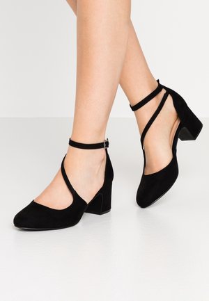 Vrouw draagt zwarte suède schoenen met blokhak en enkelbandjes op een witte vloer en effen achtergrond.