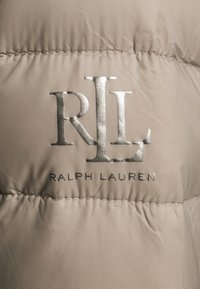 Beige vadderad jacka i tyg med sömnt detaljer. Prydd med en silverlogotyp "RLL" och texten "RALPH LAUREN" i ett slankt typsnitt.