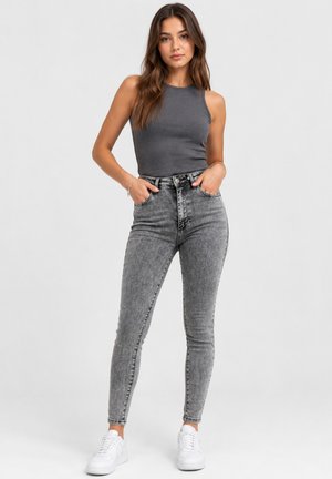Ung kvinde står med hænderne i lommerne iført en mørkegrå ærmeløs top, grå højtaljede jeans og hvide sneakers på en lys baggrund.