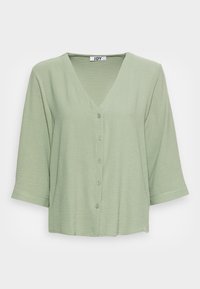 Lys grøn V-udskæring bluse med ¾-ærmer og frontknapper, lavet af tekstureret stof, præsenteret på en ensfarvet hvid baggrund.