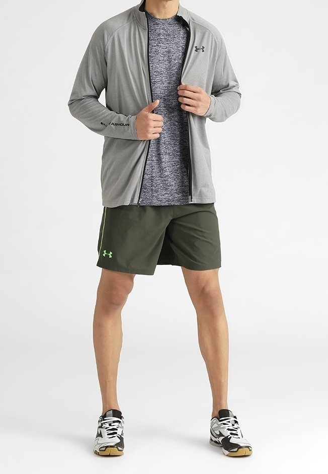 Homme portant une veste grise zippée par-dessus une chemise à motifs, un short de sport vert et des baskets noires et blanches, debout les jambes écartées.