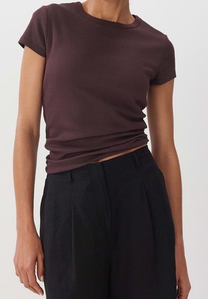 Persoon draagt een donkerbruine geribde nauwsluitende T-shirt met korte mouwen en losse zwarte hoog getailleerde broek tegen een effen achtergrond.