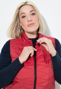 Rode gewatteerde vest met hoge kraag, ritssluiting aan de voorkant en zwarte rits, gedragen over een marineblauwe trui. Glad oppervlak, modern ontwerp.