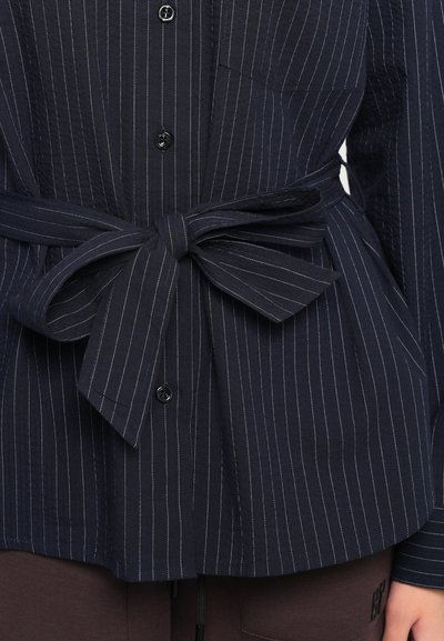 Navy striped blouse with a tie belt at the waist, featuring textured fabric and front buttons, complemented by a single pocket detail.