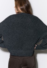 Tmavosivý, nadrozmerný pletený sveter s textúrovaným povrchom, širokými rukávmi a rebrovaným lemom, nosený cez hnedé nohavice. Zobrazený pohľad zozadu.