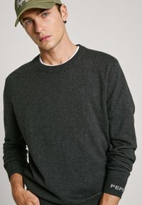 Tmavě šedý pletený svetr s kulatým výstřhem a dlouhými rukávy. Obsahuje pružné manžety a jemné logo na zápěstí. Nosený přes bílé tričko.