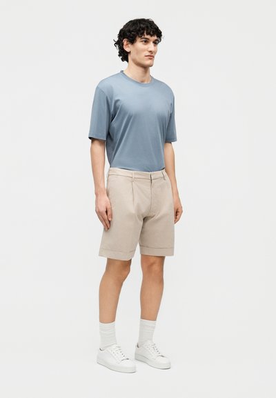 Jeune homme debout aux cheveux bouclés, portant une chemise à manches courtes bleu clair, un short beige, des chaussettes blanches et des baskets blanches, sur fond blanc.