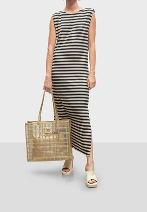 Femme portant une longue robe sans manches rayée noire et beige, tenant un grand sac cabas tissé doré, et portant des sandales beiges à plateforme à bouts ouverts.