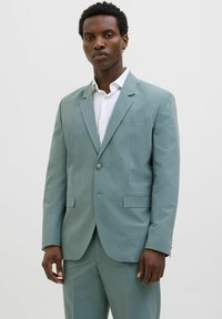 Costum teal deschis dintr-un material texturat, cu închidere printr-un singur nasture, două buzunare frontale și rever ascuțit. Cămașă albă dedesubt.