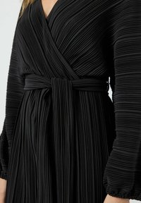 Vestido negro de pliegues con diseño cruzado, que cuenta con un lazo en la cintura y mangas largas con puños fruncidos. Tejido suave y texturizado.