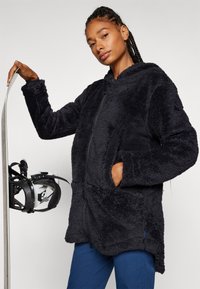 Schwarzer, weicher Fleece-Hoodie mit Reißverschluss vorne und zwei Seitentaschen, getragen mit blauen Hosen. Verfügt über einen lässigen Schnitt und eine Kapuze.