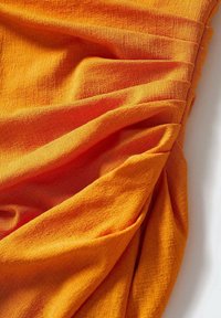 Tissu orange texturé avec un drapé doux et froissé, présentant des motifs subtils. Comprend des fronces et des plis le long d'un bord.