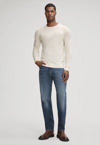 Sweter z długim rękawem w kolorze kremowym, wykonany na drutach, w połączeniu z niebieskimi dżinsami; model ma na sobie brązowe buty. Sweter ma ściągacze na rękawach, gładką teksturę i luźny krój.