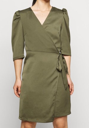 Kobieta ubrana w oliwkową sukienkę o długości do kolan, z rękawami o długości do łokcia zakończonymi bufkami oraz zawiązanym kokardą bokiem.