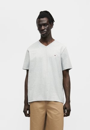 Άνδρας με σκούρο δέρμα και dreadlocks, φοράει ανοιχτογρήγορο V-λαιμό μπλουζάκι και μπεζ παντελόνια, στέκεται μπροστά σε απλό λευκό φόντο.