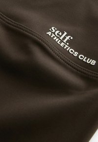 Pruun sportkangas, millel on sile pind, ja valge trükitud tekst "self. ATHLETICS CLUB" õmbluse ääres.