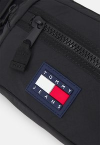 Sac en tissu noir avec une poche zippée, et un logo rectangulaire avec des blocs de couleur bleue, blanche et rouge portant l'inscription "TOMMY JEANS." Fini texturé.