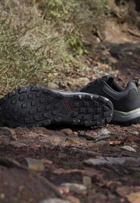 Chaussure de randonnée noire avec semelle à crampons profonds portant l'étiquette "TRAXION" posée sur un sentier rocheux en plein air entouré de verdure.