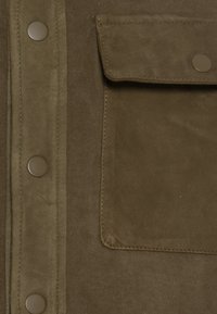 Olivgrön mocka med synliga sömmar och en lockficka, säkrad med en metallknapp. Mjuk textur med en matt yta.