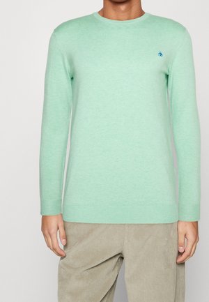 Mand iført mintgrøn langærmet sweater med lille blåt broderet logo på brystet og beige kordfløjlsbukser.