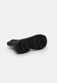 Suela de bota de goma negra con una superficie texturizada, tacos pronunciados y ranuras. El tacón tiene un contorno para mayor estabilidad.
