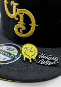 Musta hattu kultakirjonnalla, jossa on monimutkaisia kirjaimia. Mukana värikkäitä pinnejä: keltainen hymyilevä naama, jossa lukee "NL", sekä musta käsinkirjoitettu logo "Naughty League".