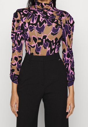 Femme portant un haut à col roulé à imprimé abstrait marron, violet et noir avec manches longues, assorti à un pantalon noir taille haute.