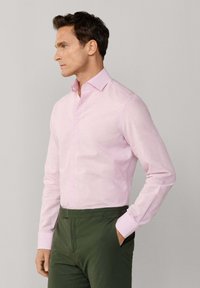 Mand iført lyserød, stribet skjorte med lange ærmer og knapper, stoppet ned i mørkegrønne bukser, stående med den ene hånd i lommen, vendt mod venstre.