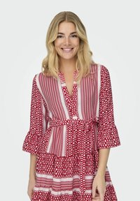 Robe à motifs rouges et blancs avec un col, des manches évasées et une taille cintrée, présentant des motifs géométriques et un ourlet en couches.