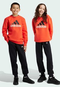 Sudaderas rojas de Adidas con logo negro, combinadas con pantalones jogger negros y zapatillas negras. Ambas prendas tienen un corte relajado y puños acanalados.