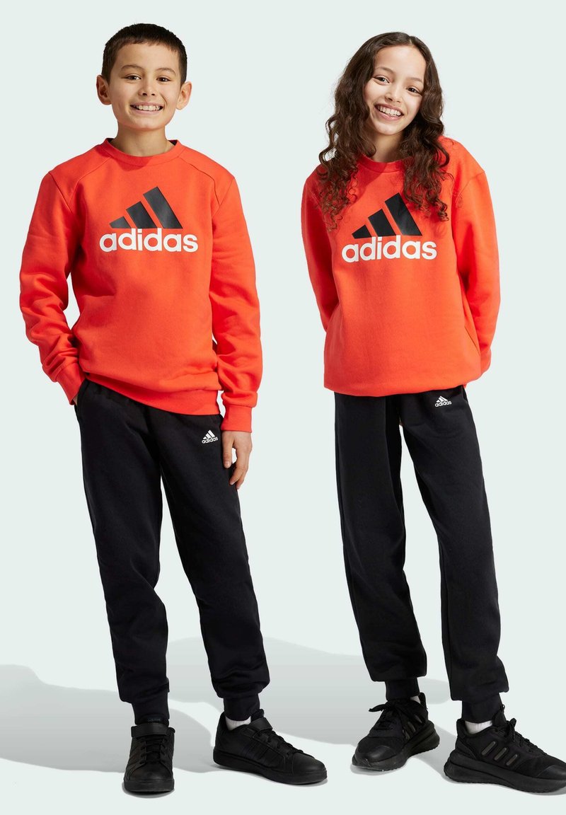 Sudaderas rojas de Adidas con logo negro, combinadas con pantalones jogger negros y zapatillas negras. Ambas prendas tienen un corte relajado y puños acanalados.