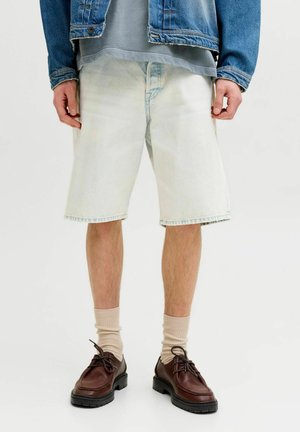 Mann iført lyseblå denimshorts, beige sokker, mørkebrune snøresko, grå skjorte og blå denimjakke som står mot hvit bakgrunn.