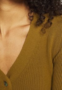 Olivgrön ribbad cardigan med V-ringning, som har knappar och ett strukturerat mönster runt halsringningen, vilket framhäver det mjuka, stickade tyget.