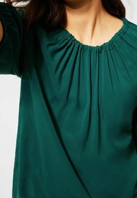 Blusa verde con scollo rotondo, dettagli arricciati e maniche ampie. Tessuto morbido con una consistenza liscia e un design fluente.