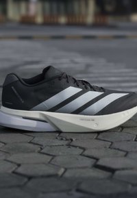 Chaussure de running Adidas Adizero Boston 13 noire et blanche avec une semelle Lightstrike Pro sur une surface pavée grise.