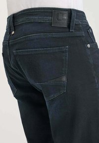 Jean en denim foncé avec une poche arrière au design angulaire distinctif, une légère décoloration et un patch en cuir texturé au niveau de la taille.