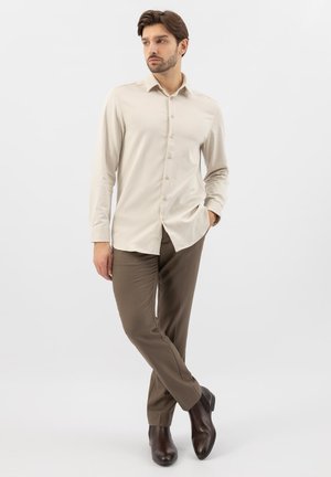 Homme portant une chemise beige à manches longues boutonnée, un pantalon marron et des chaussures habillées marron foncé, debout avec une main dans la poche, jambes croisées.